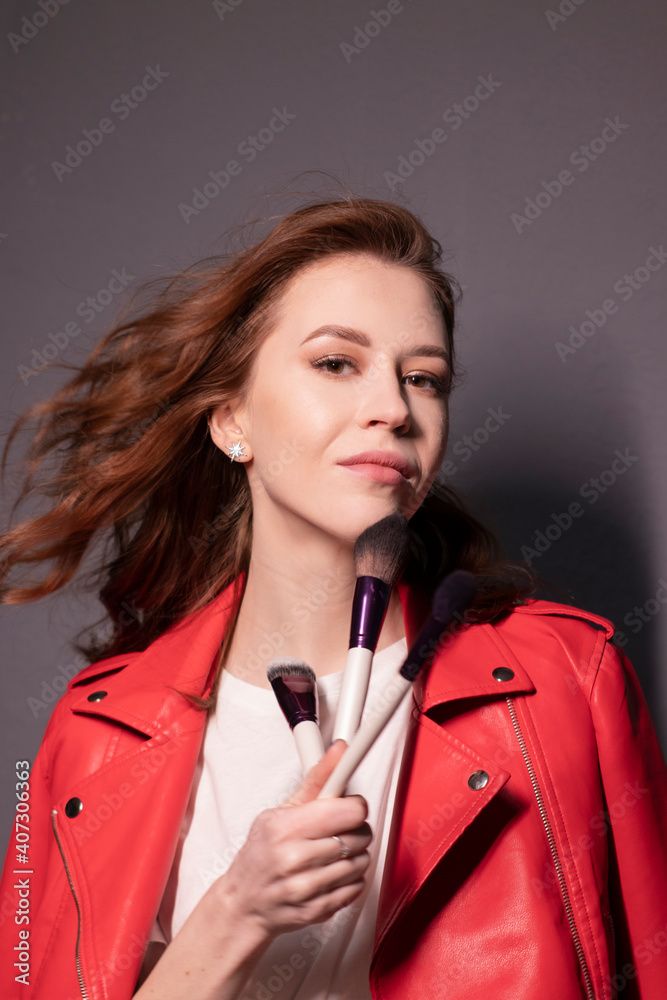 charming brunette woman in red leather jacket with makeup brushes isolated on grey wall background. professional makeup artist charming brunette woman in red leather jacket with makeup brushes isolated on grey wall background. professional makeup artist