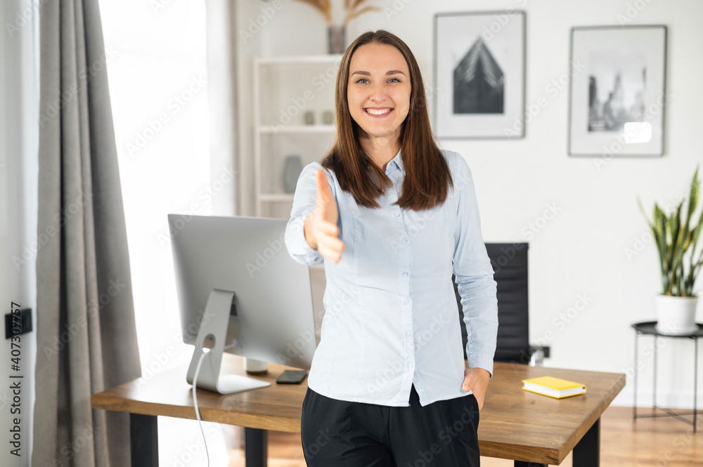 Young pleasant woman wearing smart casual shirt holds out her hand in handshake gesture, greeting, sign of good deal standing in a contemporary office space, hiring concept Young pleasant woman wearing smart casual shirt holds out her hand in handshake gesture, greeting, sign of good deal standing in a contemporary office space, hiring concept
