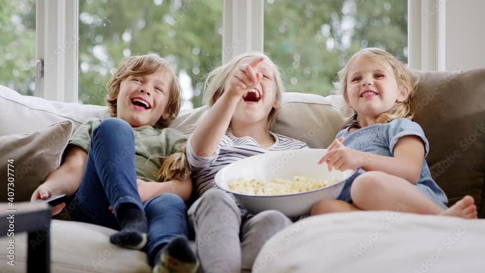 Three children sitting on sofa at home laughing and watching TV with popcorn together - shot in slow motion Three children sitting on sofa at home laughing and watching TV with popcorn together - shot in slow motion