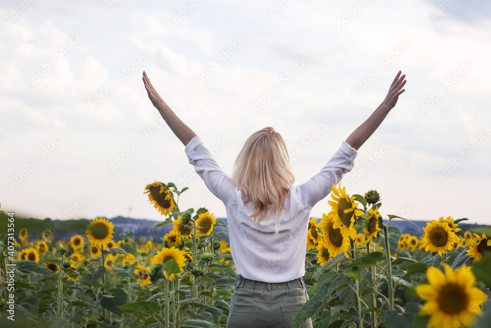 Happy adult blonde girl in a white shirt and green jeans in the middle of a field of yellow sunflowers. Young woman with hands raised up