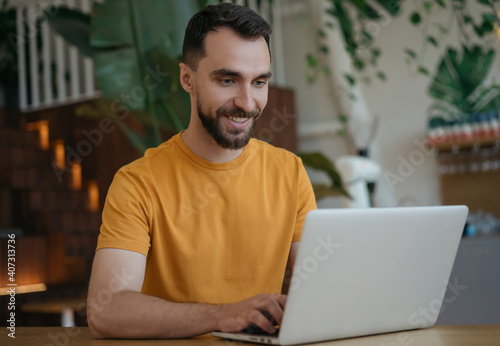 Attractive smiling man using laptop computer, working from home. Portrait of young freelancer copywriter typing, sitting at workplace. Student studying, distance learning, searching, online education © ARUTA Images