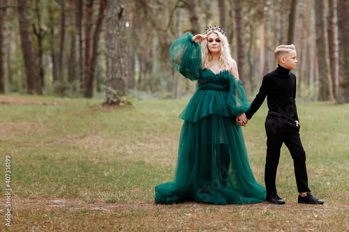 young beautiful blonde woman queen with young boy in black outfit. Princess mother walks with son. autumn green forest mystic. Vintage medieval shiny crown. Long evening green dress. magic fantasy