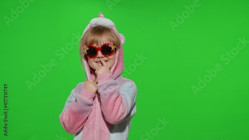 Young little blonde child smiling, puts on sunglasses, fooling around in unicorn costume on chroma key green background. Portrait of kid girl animator in unicorn pajamas. Copy space
