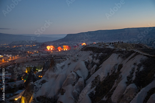 Goreme city night lights and hot air balloons glowing in early morning while inflating among rocks of mountain landscape in Cappadocia. Colorful illuminated balloons in blue hour of sunrise