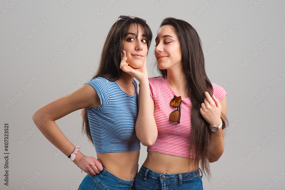 Young twin sisters. Pretty teen twins wearing striped t-shirts and jean shorts on light grey background Young twin sisters. Pretty teen twins wearing striped t-shirts and jean shorts on light grey background