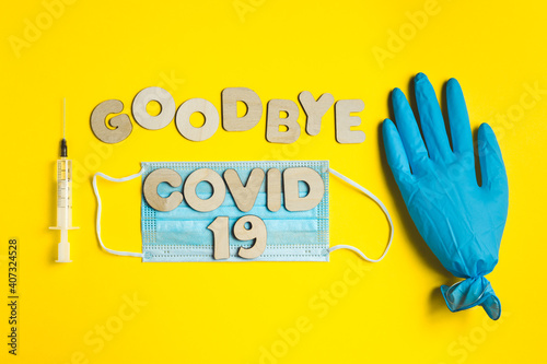 Words GOODBYE COVID 19 from wooden letters and syringe with vaccine, inflated medical glove waving bye-bye and facemask. Vaccination and the end of epidemic concept.