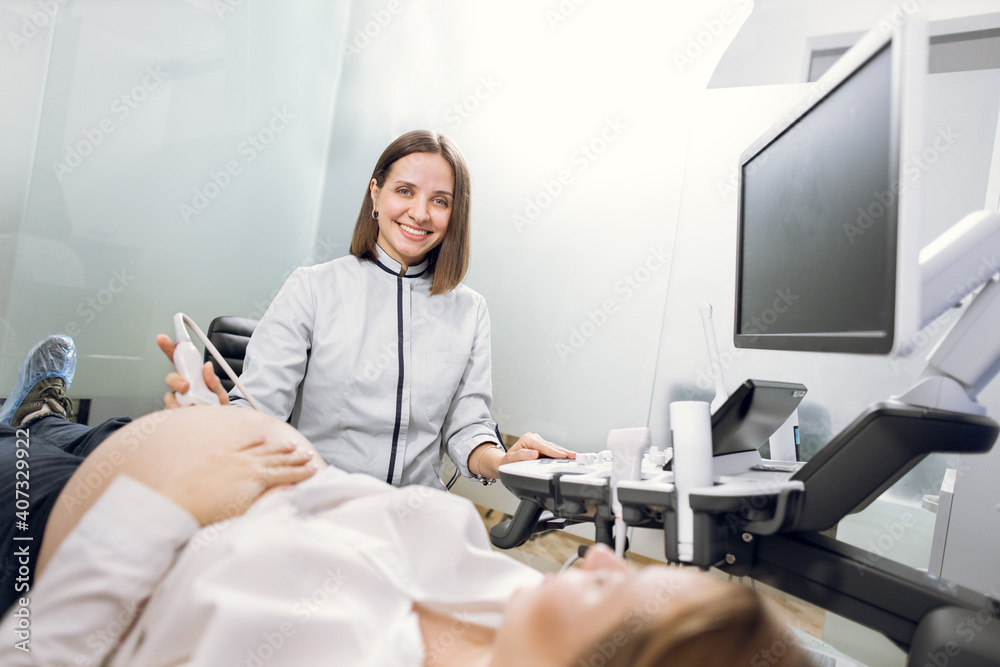 High-skilled professional attractive woman doctor doing 3d ultrasound on belly of pregnant woman in clinic, smiling to camera. Ultrasound for pregnant woman High-skilled professional attractive woman doctor doing 3d ultrasound on belly of pregnant woman in clinic, smiling to camera. Ultrasound for pregnant woman