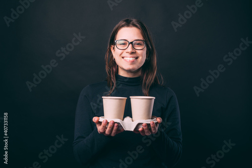 Joyful young woman over dark background holding two cups of coffee to take away.