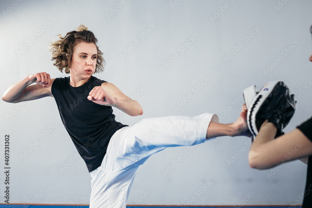 Close-up of barefoot woman leg practicing kicking with taekwondo coach who holds boxing paw. Concentrated active female kicking boxing shield with personal coach at gym. Fighters training together
