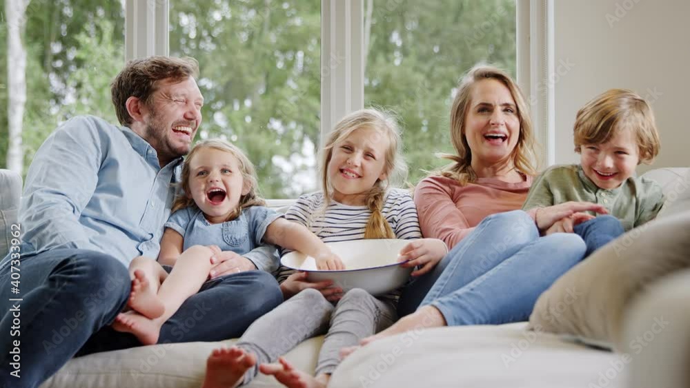 Family sitting on sofa at home laughing and watching TV with popcorn together - shot in slow motion Family sitting on sofa at home laughing and watching TV with popcorn together - shot in slow motion