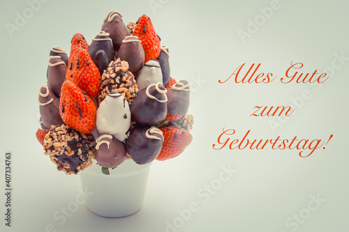 Happy birthday greeting card in German that reads Alles Gute zum Geburtstag with red lettering; A bundle of edible flowers, arrangement of strawberries covered with chocolate isolated on white