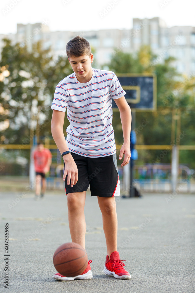 Young cute boy plays basketball at the outdoor streetball court on a sunny summer day. Teenager player in action dribbling the ball. Hobby, active lifestyle, sports for kids. Young cute boy plays basketball at the outdoor streetball court on a sunny summer day. Teenager player in action dribbling the ball. Hobby, active lifestyle, sports for kids.