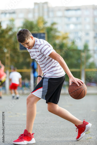Young cute boy plays basketball at the outdoor streetball court on a sunny summer day. Teenager player in action dribbling the ball. Hobby, active lifestyle, sports for kids. © Natali