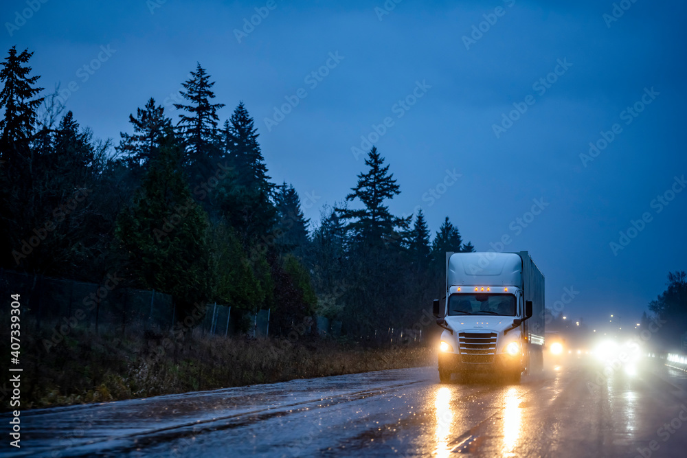 Big rig white semi truck with turned on head lights transporting cargo in semi trailer running on the night dark wet road with reflection in rain weather