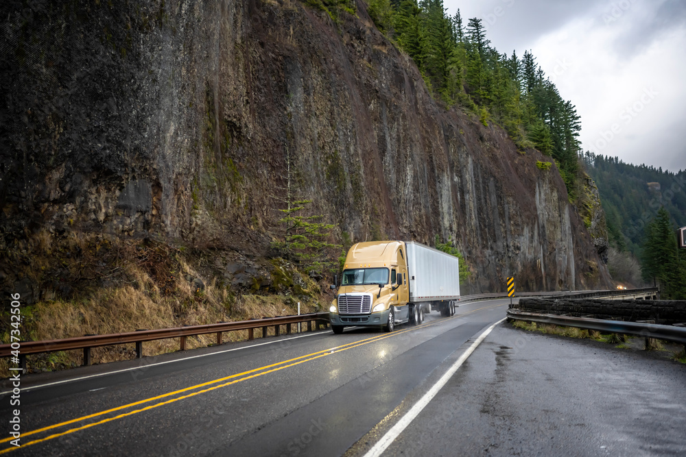 Wet yellow big rig semi truck transporting cargo in dry van semi trailer running on the mountain winding slippery road with rock cliff wall on the side at raining weather