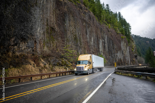 Wet yellow big rig semi truck transporting cargo in dry van semi trailer running on the mountain winding slippery road with rock cliff wall on the side at raining weather