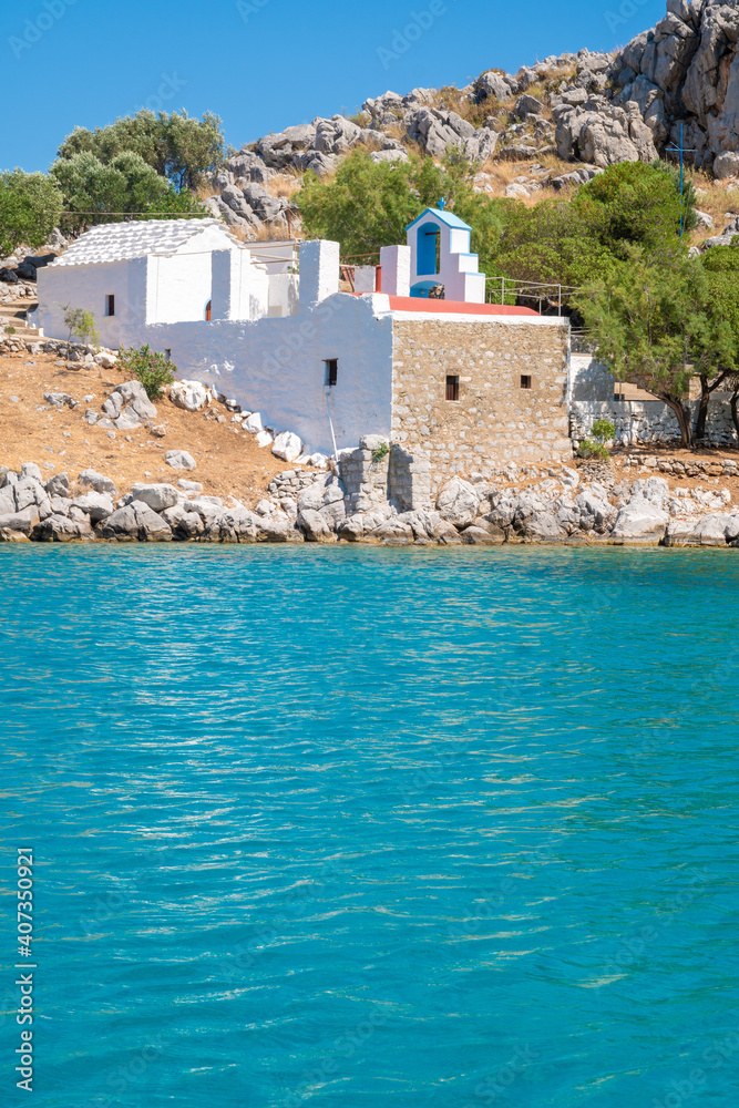 View from the sea into a bay with a stone building between rocks. A Greek Orthodox church in white and blue in harmony with the nature and turquoise calm waters