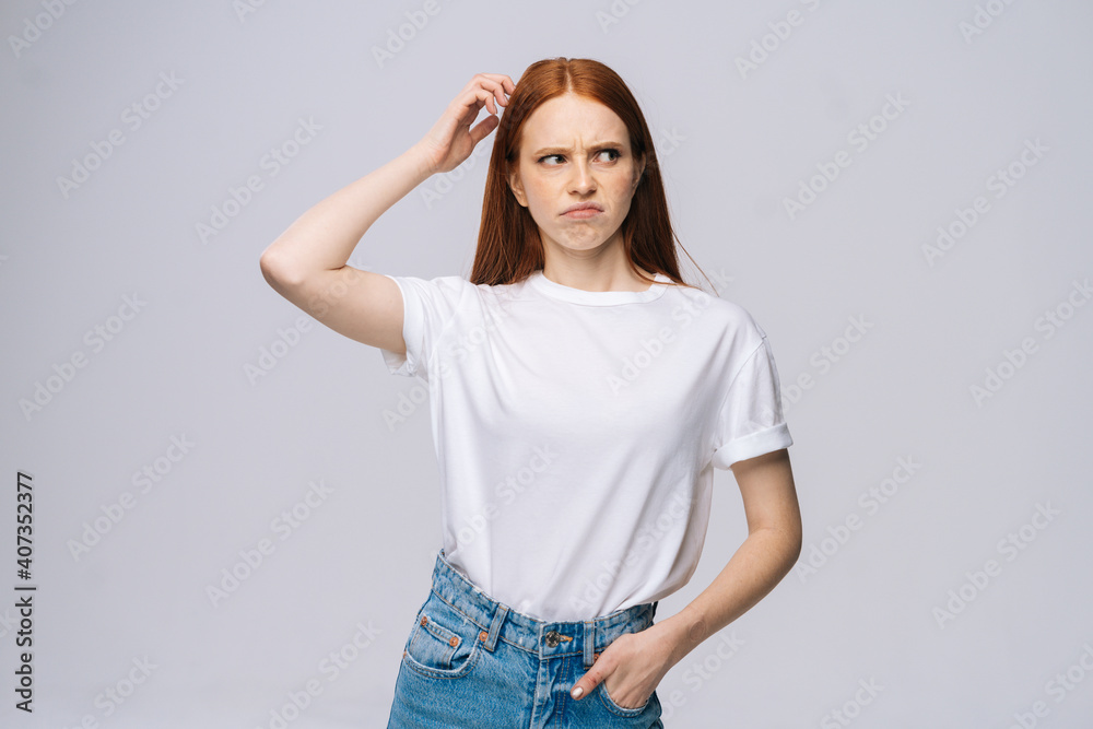 Astonished confused young woman wearing T-shirt and denim pants touching head with hand on isolated white background. Pretty lady model with red hair emotionally showing facial expressions. Astonished confused young woman wearing T-shirt and denim pants touching head with hand on isolated white background. Pretty lady model with red hair emotionally showing facial expressions.
