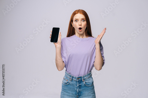 Shocked young woman wearing stylish casual clothes holding cell phone with black empty mobile screen. Pretty lady model with red hair emotionally showing facial expressions in studio, copyspace.