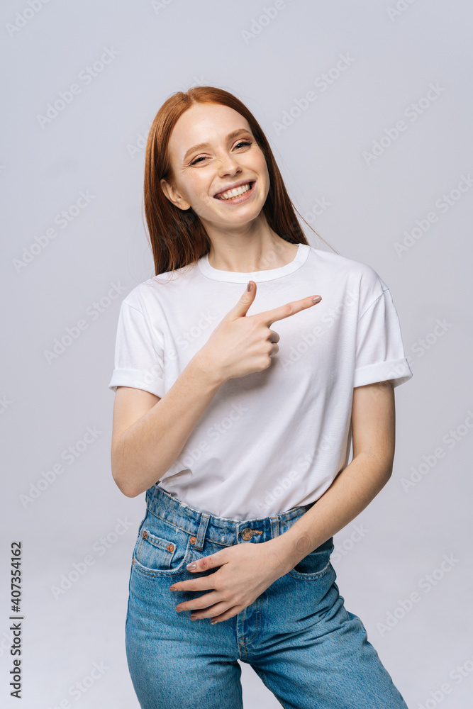 Cheerful young woman wearing T-shirt and denim pants pointing with finger at blank copy space aside on isolated white background. Pretty lady model with red hair emotionally showing facial expressions Cheerful young woman wearing T-shirt and denim pants pointing with finger at blank copy space aside on isolated white background. Pretty lady model with red hair emotionally showing facial expressions