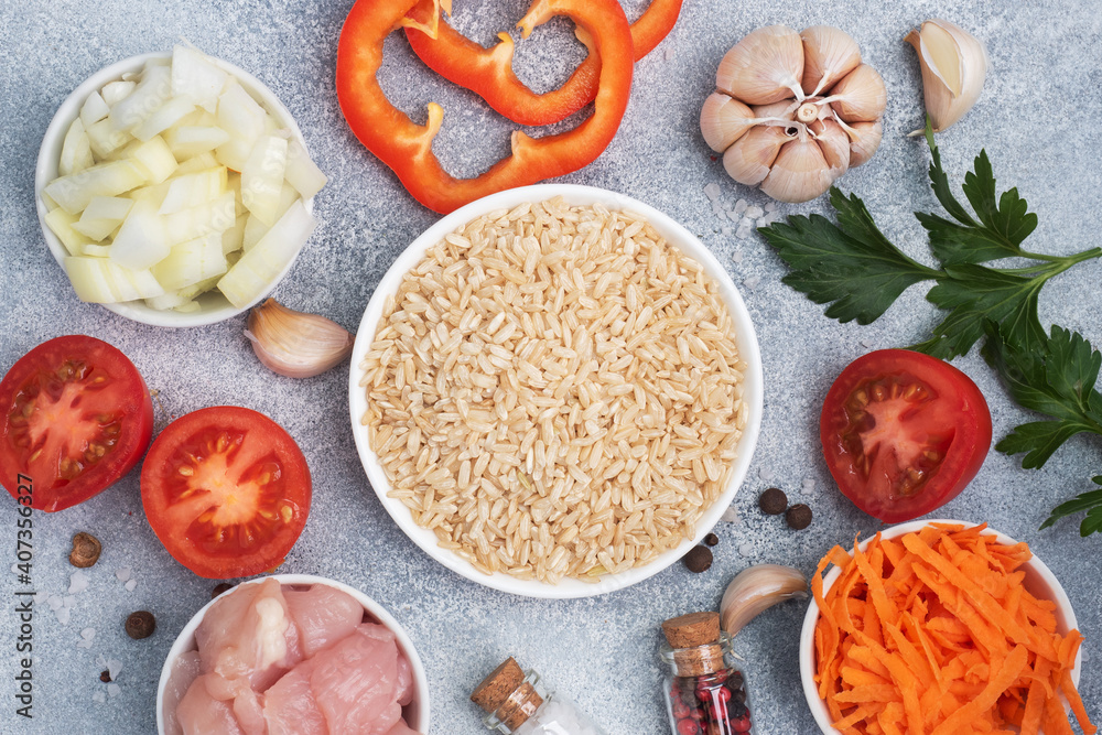 Raw ingredients for cooking pilaf, Brown rice and chicken fillet, grated carrots and chopped onions, pepper tomato garlic herbs and spices. Gray background, top view. Raw ingredients for cooking pilaf, Brown rice and chicken fillet, grated carrots and chopped onions, pepper tomato garlic herbs and spices. Gray background, top view.