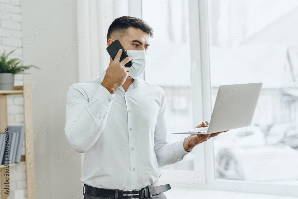 Handsome man businessman in medical mask using his laptop while talking on his cellphone Handsome man businessman in medical mask using his laptop while talking on his cellphone