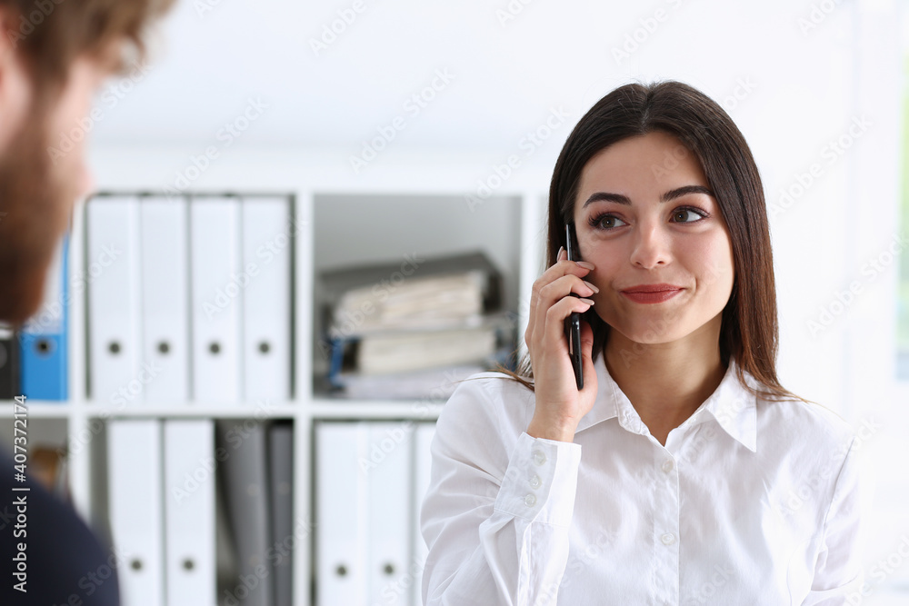 Beautiful businesswoman brunette portrait in an office in a white shirt is holding a phone and talking to a customer remotely looking into the distance listening to what they say and receiving