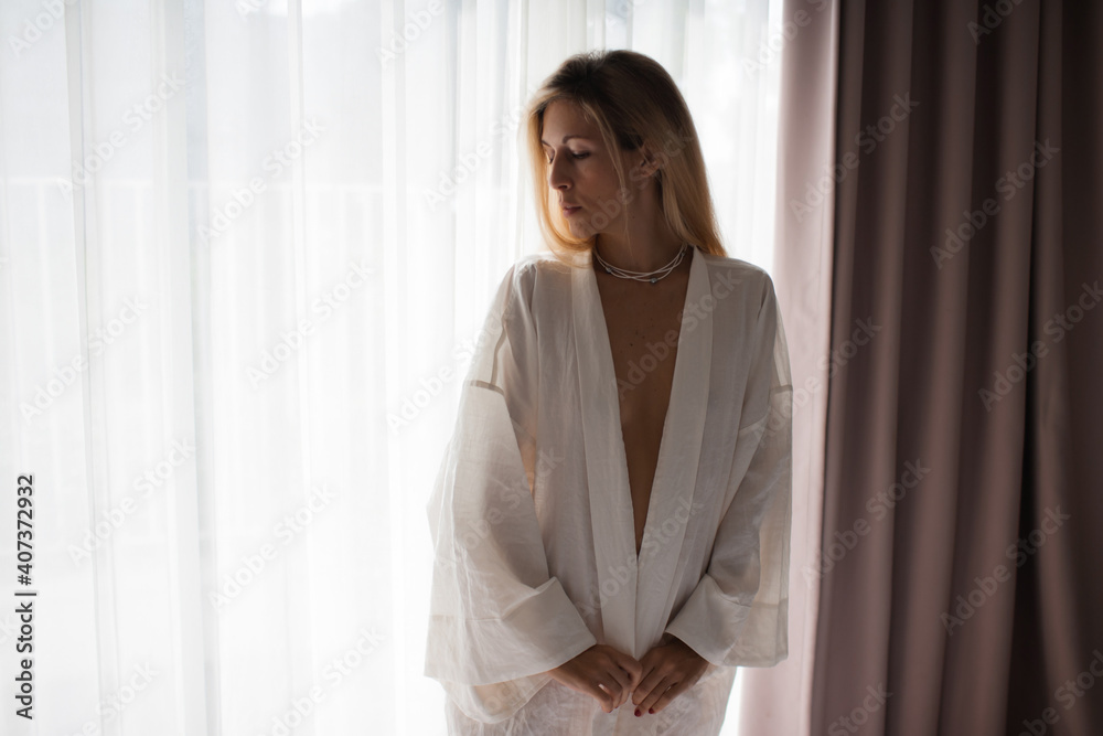 Beautiful blond woman is enjoying morning at home in white handmade linen kimono robe wearing elegant pearl jewelry next to the window Beautiful blond woman is enjoying morning at home in white handmade linen kimono robe wearing elegant pearl jewelry next to the window