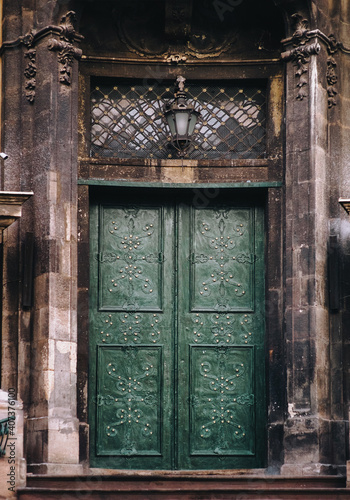 An old medieval green wooden door on the facade of the Dominican Cathedral in the Baroque style in Lviv is decorated with decorative wrought iron patterns. Lamp and window grill above the doorway.