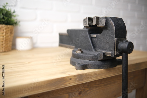 Close-up of a black vise with a screw grip and a cast-iron frame mounted on a wooden countertop, in a machine shop. The white brick background is blurred