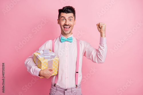 Photo portrait of cheerful man with raised fist holding gift box isolated on pastel pink colored background