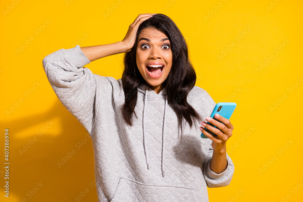 Photo of funky impressed dark skin woman wear grey hoodie holding modern device arm head isolated yellow color background Photo of funky impressed dark skin woman wear grey hoodie holding modern device arm head isolated yellow color background
