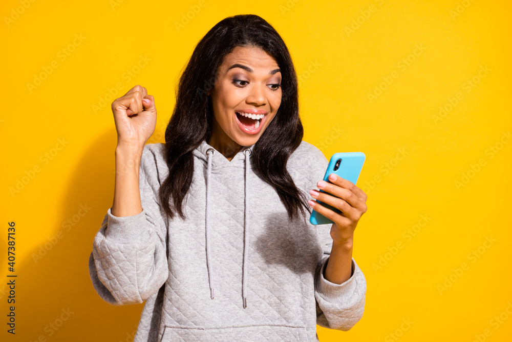 Photo of impressed lucky dark skin woman wear grey hoodie looking modern device isolated yellow color background Photo of impressed lucky dark skin woman wear grey hoodie looking modern device isolated yellow color background