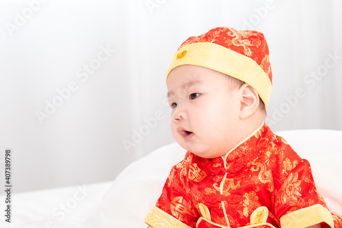 Adorable Asian boy in red Chinese Costume clothes sitting on bed at home looking for somethin with curiosity, innocent cute baby feel sleepy yawning boring waiting for mother feeding photo