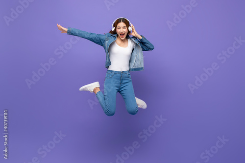 Fun energetic young Asian woman jumping and listening to music on headphones isolated on purple studio background