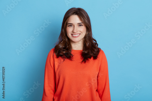 Positive joyful young beautiful female with dark hair wearing casual clothing, looking at camera and pleasantly smiling, wearing orange sweater, standing isolated over blue background.