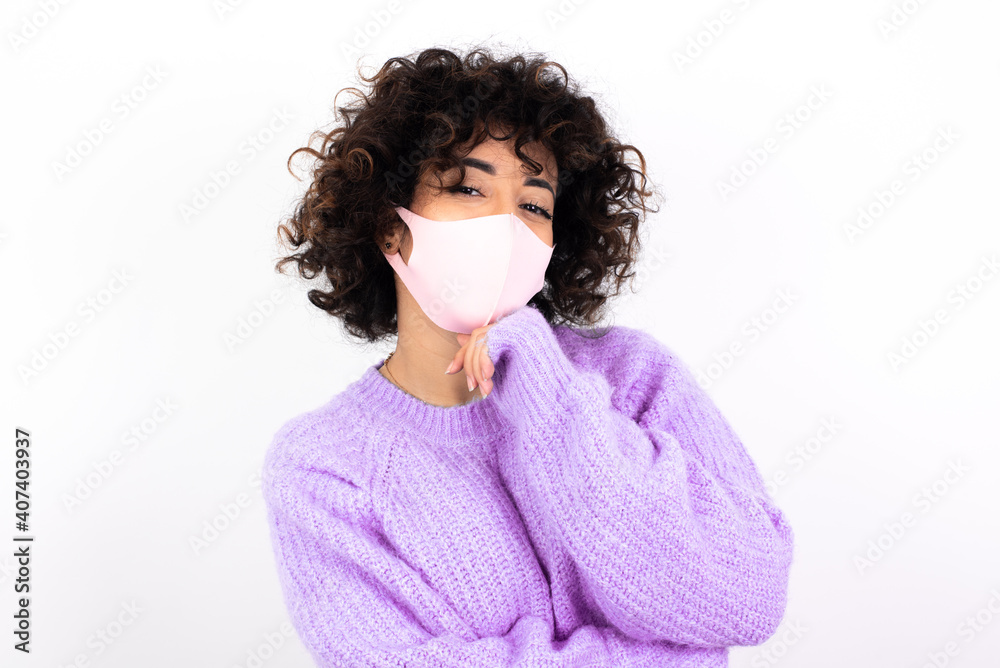Optimistic young beautiful caucasian woman wearing medical mask standing against white wall keeps hands partly crossed and hand under chin, looks at camera with pleasure. Happy emotions concept.