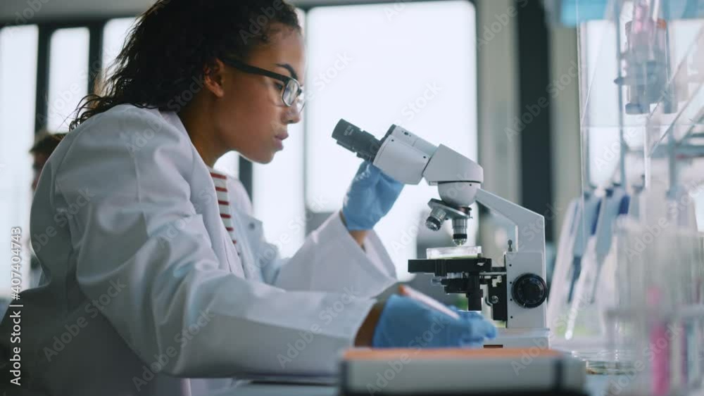 Medical Science Laboratory: Beautiful Black Scientist Looking Under Microscope, writes down Analysis of Test. Young Biotechnology Specialist, Using Advanced Equipment. Side View Zoom In Portrait Medical Science Laboratory: Beautiful Black Scientist Looking Under Microscope, writes down Analysis of Test. Young Biotechnology Specialist, Using Advanced Equipment. Side View Zoom In Portrait