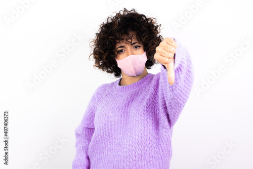 young beautiful caucasian woman wearing medical mask standing against white wall feeling angry, annoyed, disappointed or displeased, showing thumbs down with a serious look © Roquillo