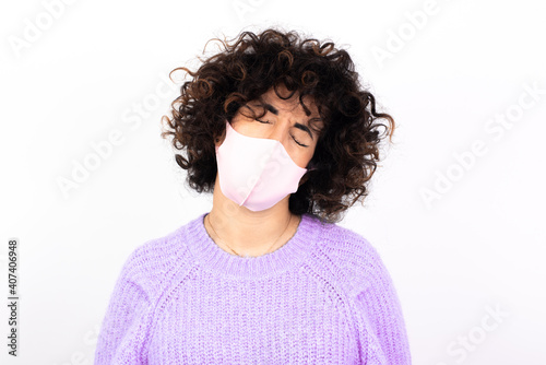 Dismal gloomy rejected young beautiful caucasian woman wearing medical mask standing against white wall has problems and difficulties, curves lower lip and closes eyes in despair, being in depression