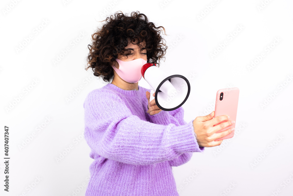 Crazy impressed young beautiful caucasian woman wearing medical mask standing against white wall use cell phone reads incredible news on social media information stare, screams wow omg. Crazy impressed young beautiful caucasian woman wearing medical mask standing against white wall use cell phone reads incredible news on social media information stare, screams wow omg.