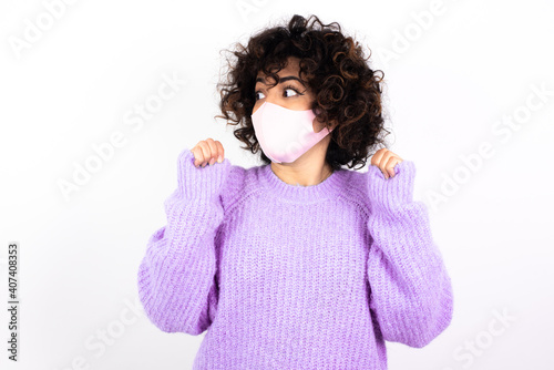 young beautiful caucasian woman wearing medical mask standing against white wall clenches fists and awaits for something nice happened looks away bites lips and waits announcement of results © Roquillo