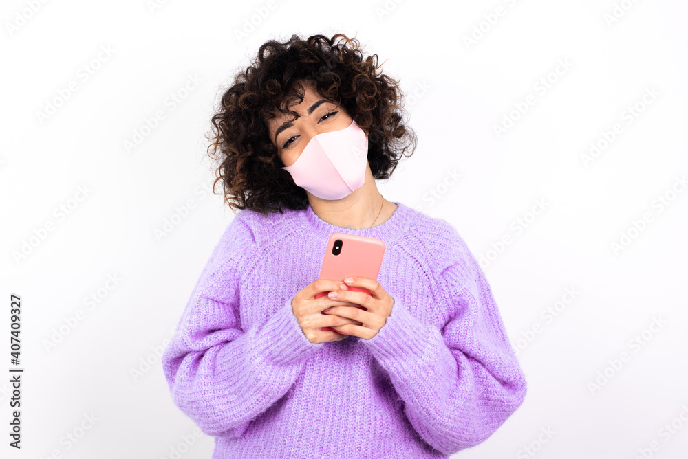young beautiful caucasian woman wearing medical mask standing against white wall Mock up copy space. Using mobile phone, typing sms message young beautiful caucasian woman wearing medical mask standing against white wall Mock up copy space. Using mobile phone, typing sms message