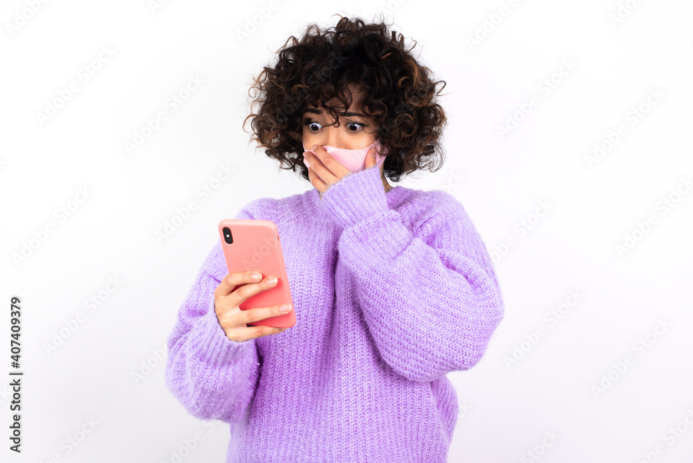 young beautiful caucasian woman wearing medical mask standing against white wall being deeply surprised, stares at smartphone display, reads shocking news on website, Omg, its horrible! young beautiful caucasian woman wearing medical mask standing against white wall being deeply surprised, stares at smartphone display, reads shocking news on website, Omg, its horrible!