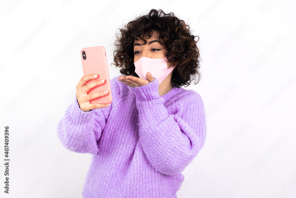 young beautiful caucasian woman wearing medical mask standing against white wall blows air kiss at camera of smartphone and takes selfie, sends mwah via online call. young beautiful caucasian woman wearing medical mask standing against white wall blows air kiss at camera of smartphone and takes selfie, sends mwah via online call.
