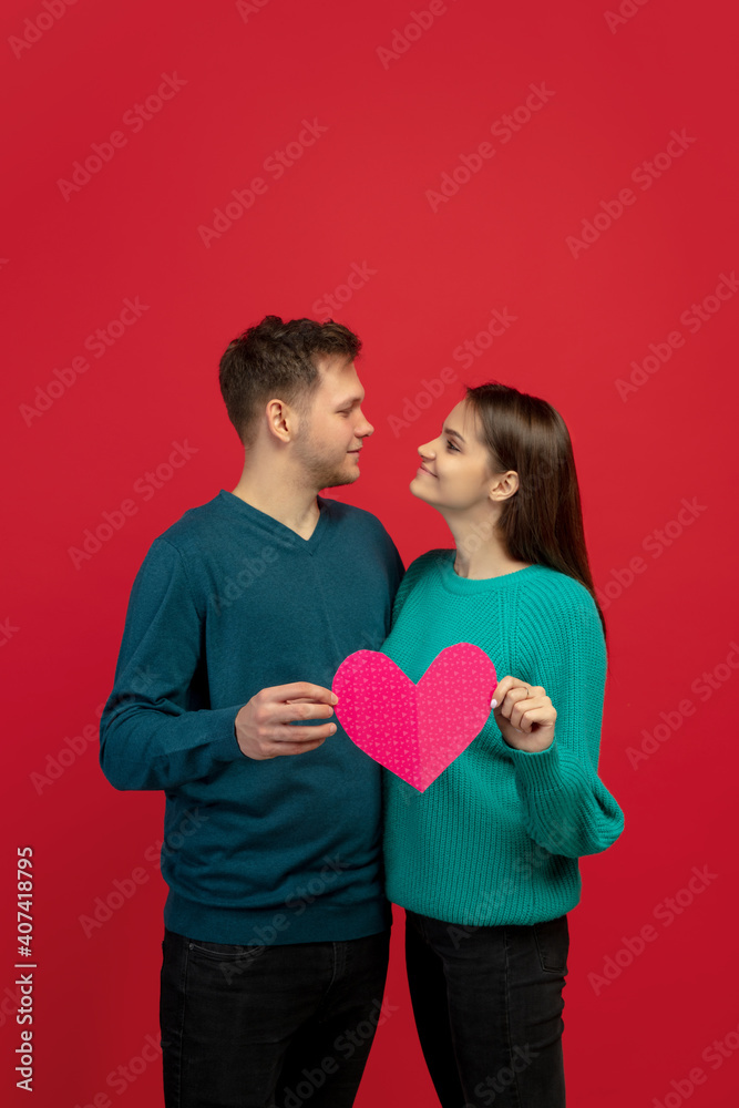 Holding pink heart. Beautiful couple in love on red studio background. Saint Valentine's Day, love, relationship and human emotions concept. Copyspace. Young man and woman look happy together.