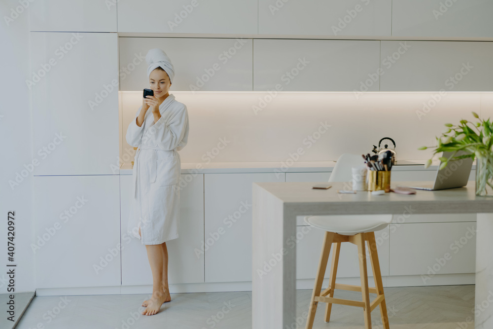 Full length shot of young beautiful woman dressed in white bathrobe applies face cream looks in mirror takes care of her skin poses near kitchen furniture stands bare feet. People beauty facial care Full length shot of young beautiful woman dressed in white bathrobe applies face cream looks in mirror takes care of her skin poses near kitchen furniture stands bare feet. People beauty facial care