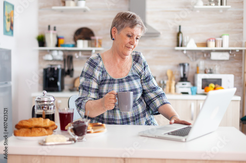 Senior businesswoman drinking coffee and working on laptop in kitchen during breakfast. Elderly retired person working from home, telecommuting using remote internet job online communication on modern