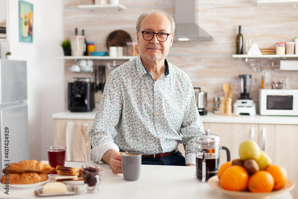 Mature man with wrinkles looking at camera in modern kitchen enjoying a cup of coffee. Portrait of relaxed elderly senior person in the morning, drinking fresh warm drink, healthy smiling adult face - obrazy, fototapety, plakaty 