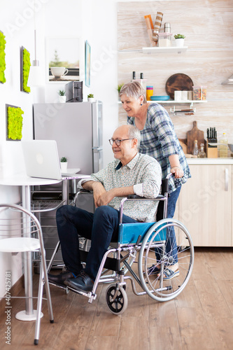 Disabled senior man in wheelchair and his wife having a video conference on laptop in kitchen. Paralysed elderly person using modern communication techonolgy, reading, searching, surfing on internet